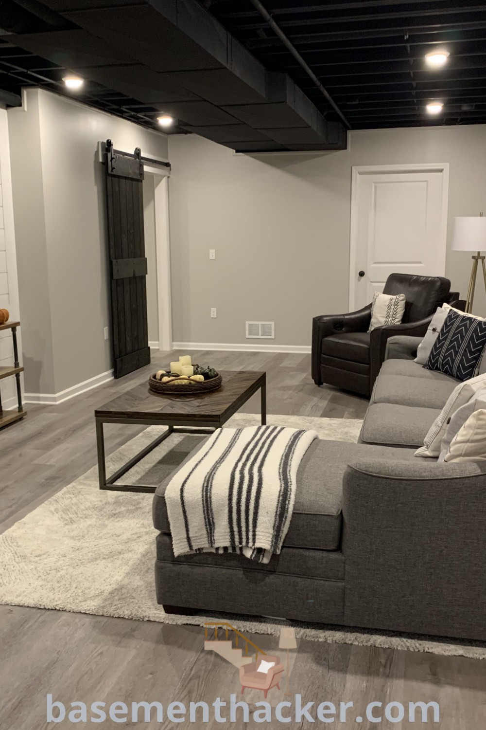 A cozy living room in a black and gray basement layout, showcasing functional furniture and a flat-screen TV mounted on the wall. This stylish bonus room offers innovative ideas for a stunning revamp and the best basement remodeling ideas for your home, featured on basementhacker.com.