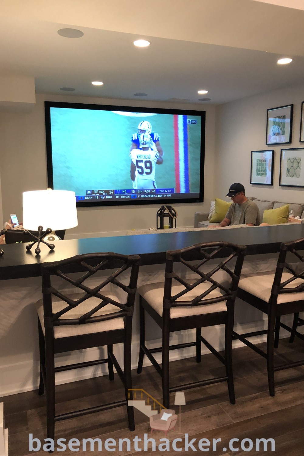 A cozy basement entertainment room with two people sitting at a bar watching TV, surrounded by chairs. This space showcases innovative ideas for designing a small finished basement, offering the best basement remodeling ideas for your home. Find more inspiration at basementhacker.com.