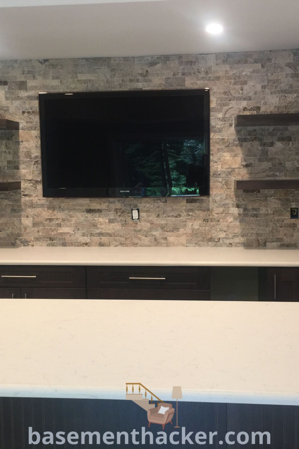 An empty kitchen with white countertops and dark wood cabinets, featuring a flat screen TV mounted on the wall, showcasing stylish basement bar kitchen ideas for ultimate entertaining. Discover more innovative ideas for designing your basement space at basementhacker.com.