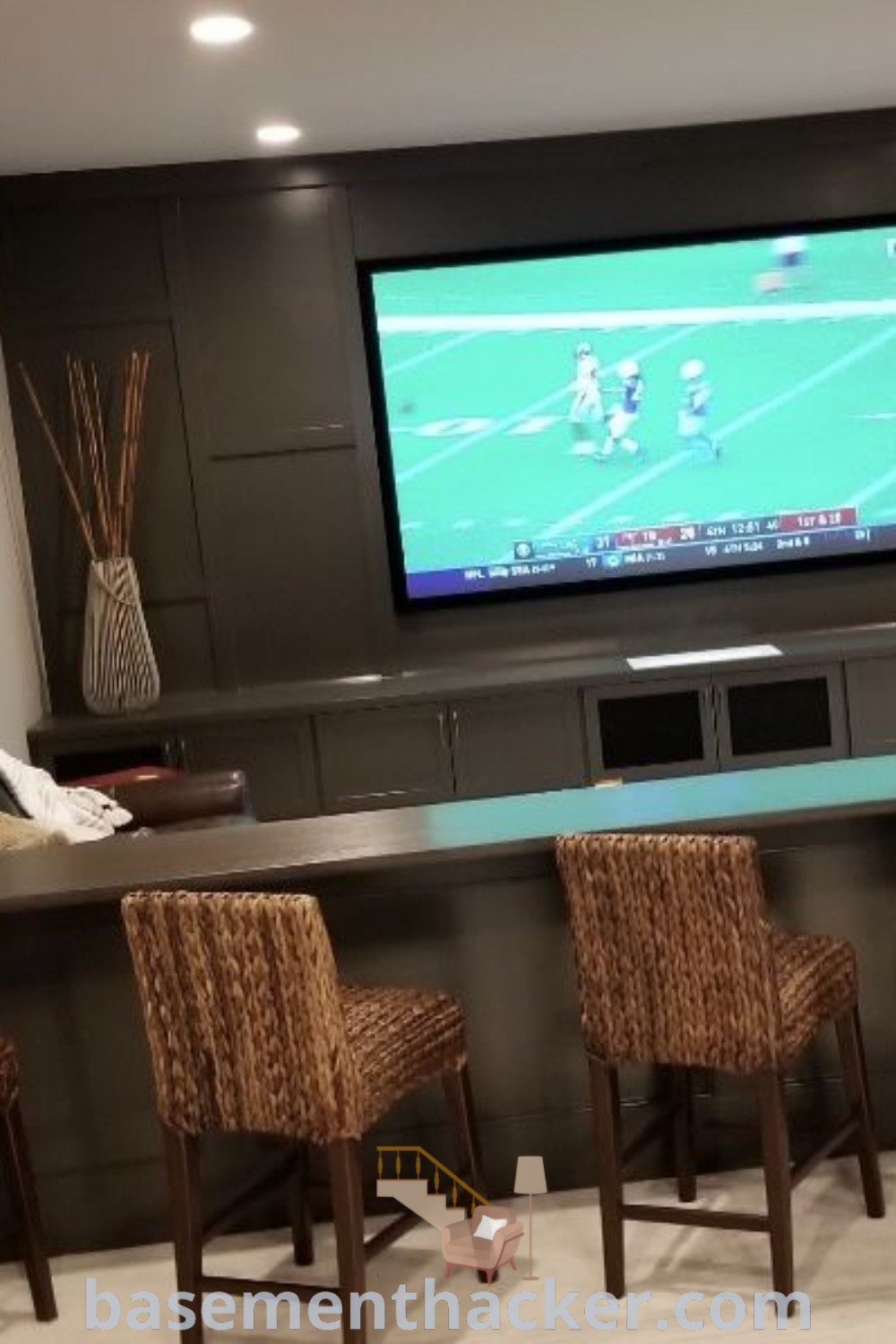 A man standing in front of a bar in a basement designed for ultimate entertaining, featuring four chairs and a television. This space embodies innovative ideas for a cozy family cave, sports bar, and multifunctional room ideas for stylish bonus spaces. Explore more at basementhacker.com.