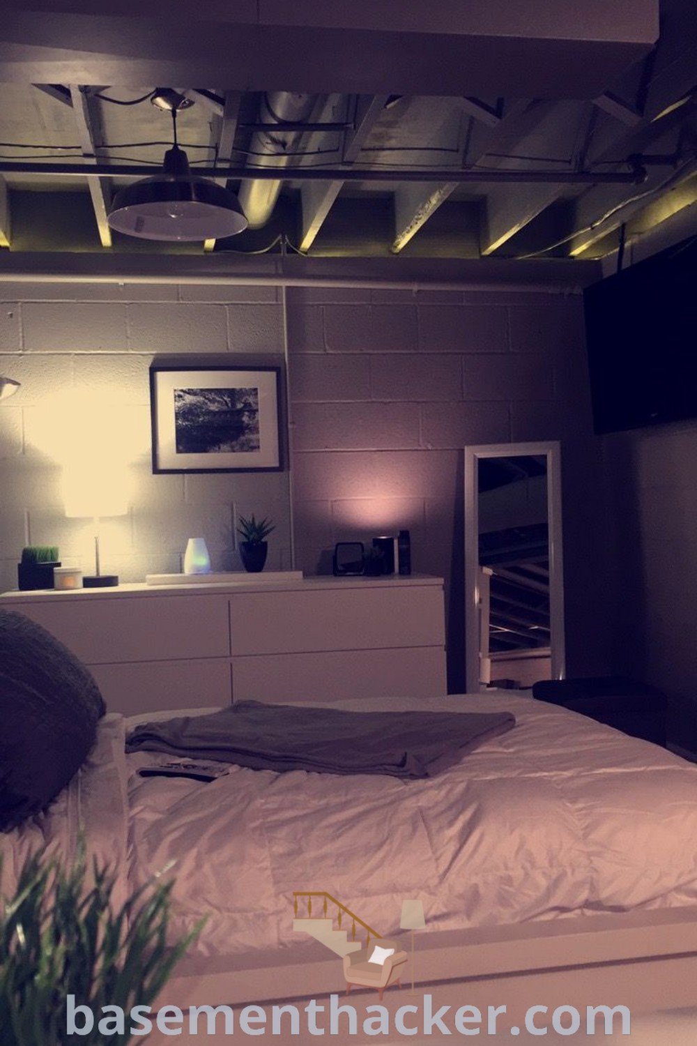 A cozy basement bedroom with a white bed under a flat-screen TV next to a plant in a vase, showcasing innovative basement makeover ideas for a stylish and functional space. Perfect for couples, this room reflects the best basement remodeling ideas, enhancing comfort and aesthetic appeal. Discover more at basementhacker.com.