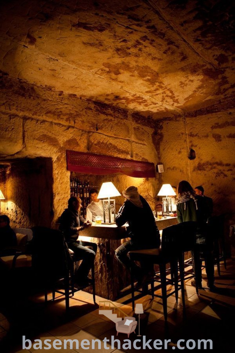 A cozy underground bar with people seated at tables, dim lighting, and lamps, creating a classic speakeasy atmosphere. Discover innovative ideas for designing your own pub interior at basementhacker.com.