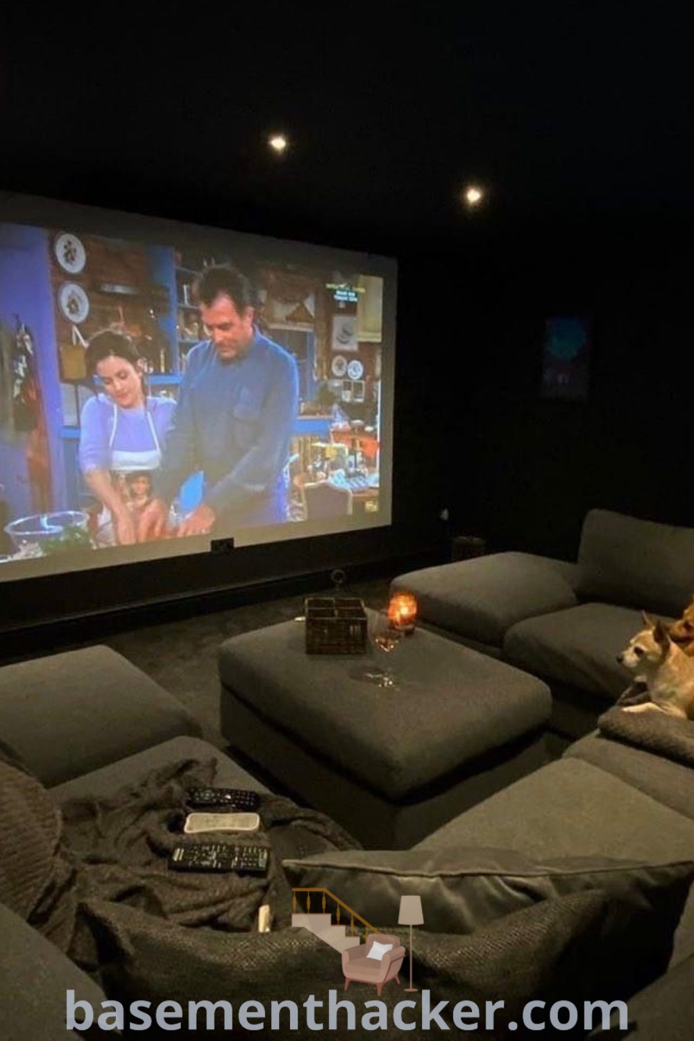 A dog laying on a couch in a home theater room in front of a large screen, with two people watching an exciting movie. This cozy setup offers innovative ideas for designing a stylish and functional basement for ultimate entertaining. Find more home cinema room ideas at basementhacker.com.