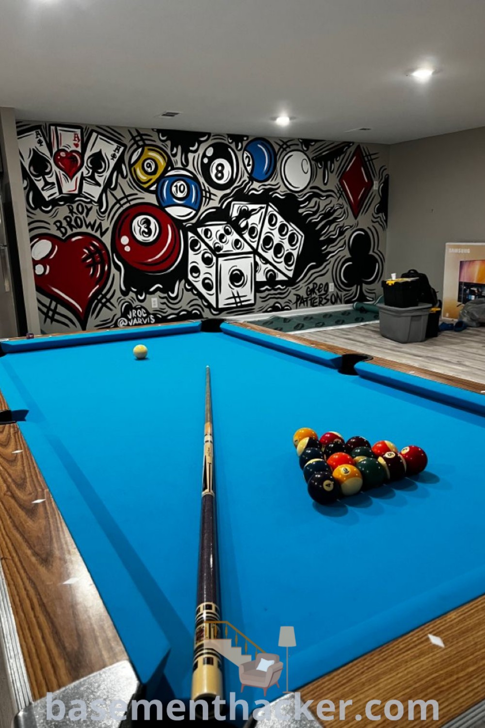A pool table with cues and balls in front of a colorful wall mural, creating a stylish game room atmosphere. This decor embodies basement makeover ideas for a captivating entertaining space, showcasing innovative designs for a cozy billiards room. For more inspiration, visit basementhacker.com.