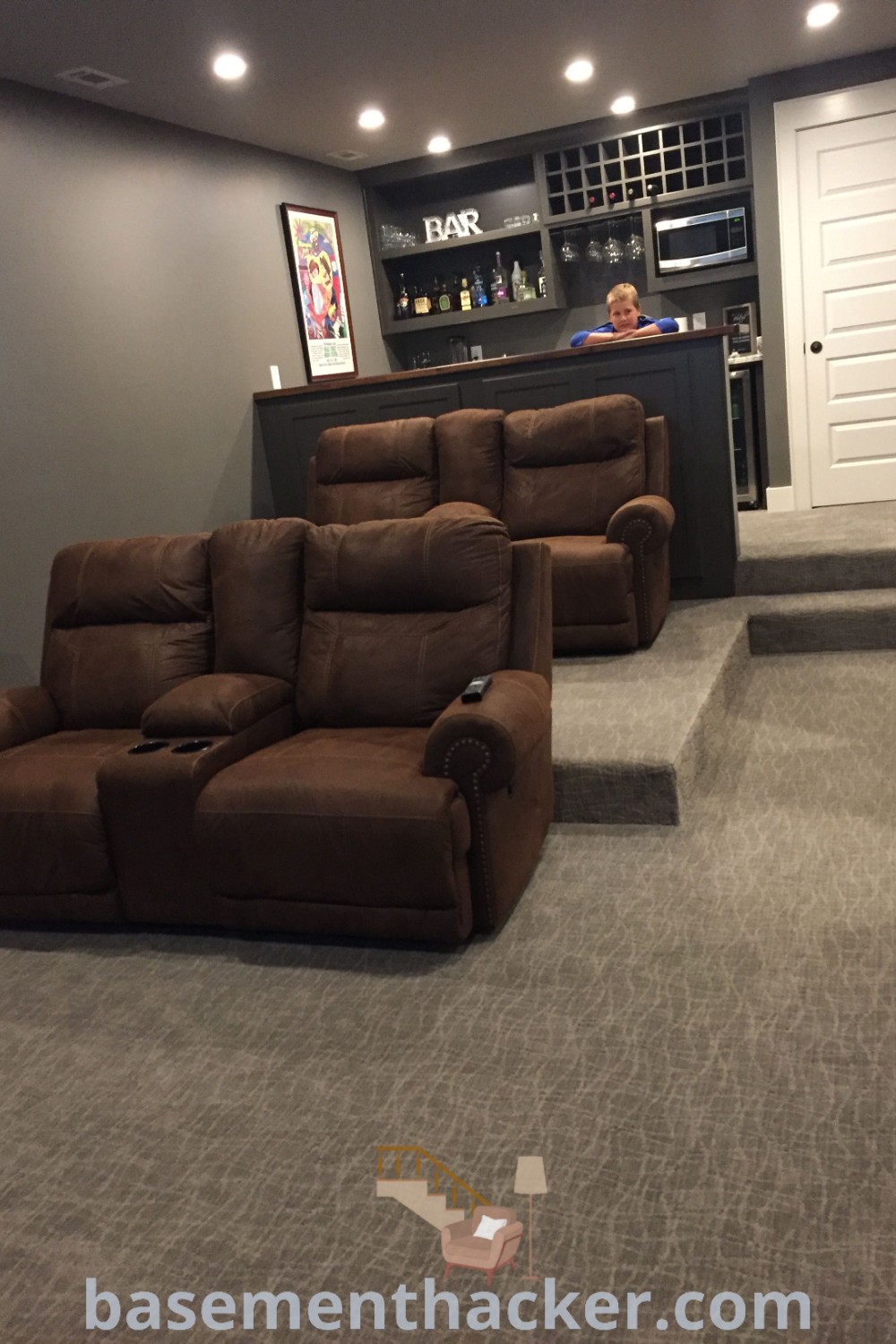 A cozy home theater room with two recliners and a couch, complemented by stairs leading up to a stylish bar, offering innovative basement makeover ideas for a stunning revamp. This setup emphasizes budget-friendly solutions while being perfect for ultimate entertaining and movie nights. Discover more ideas at basementhacker.com.