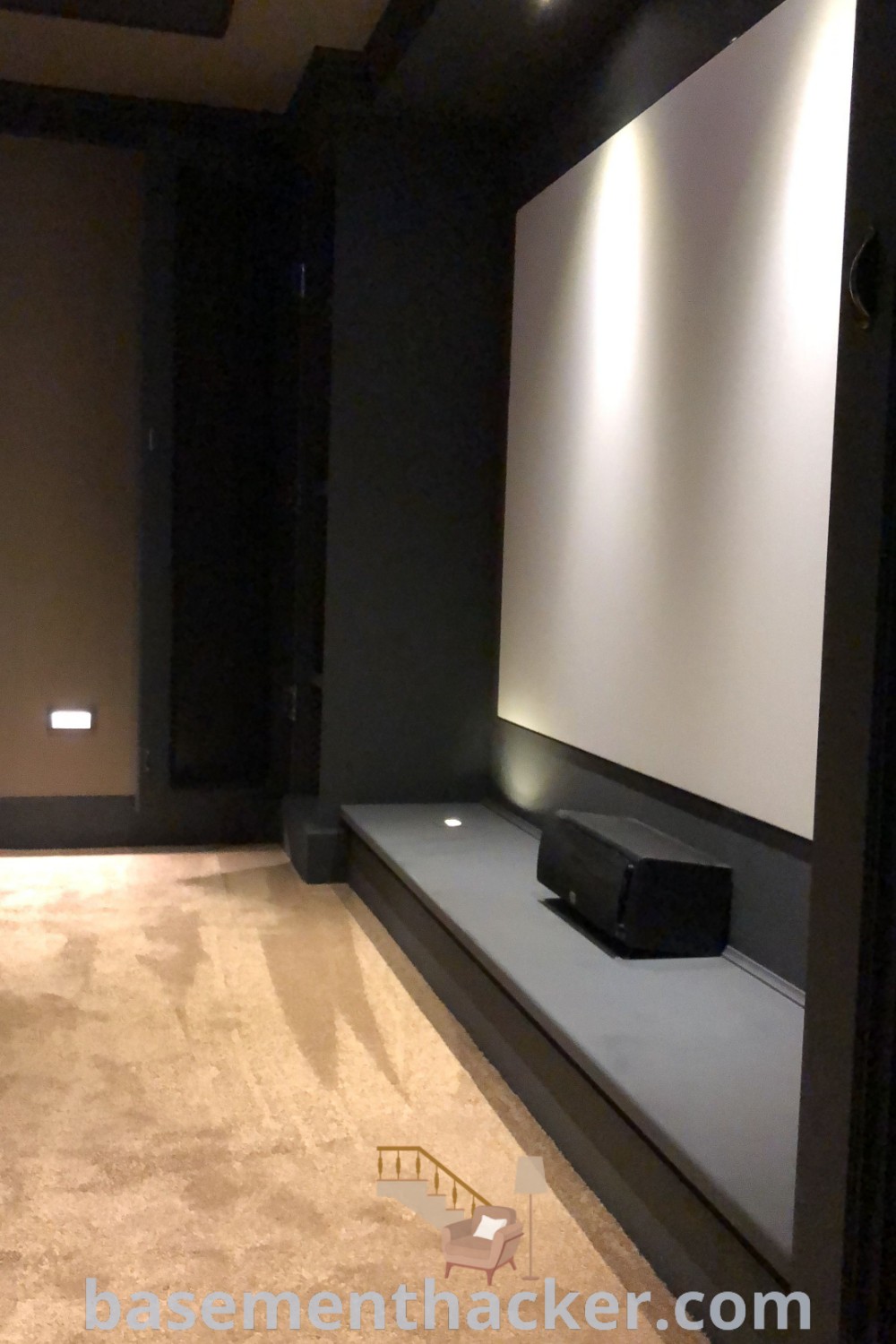 An empty room with lights on the ceiling and a projector screen, showcasing innovative ideas for a small home theater. This design is perfect for a basement movie theater and provides stylish bonus room design ideas for families. Explore more home theater room design inspirations at basementhacker.com.