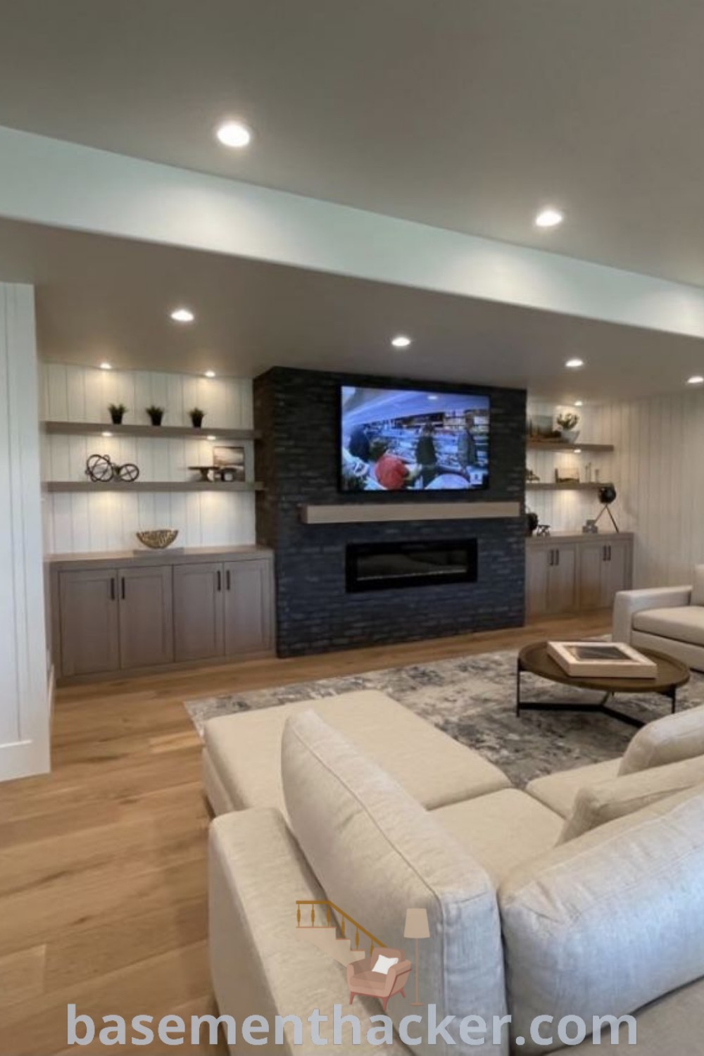A large living room in a finished basement with white couches and a flat-screen TV on the wall, showcasing basement aesthetic and design ideas for a cozy and organized home. Find innovative basement makeover ideas for stylish bonus rooms at basementhacker.com.