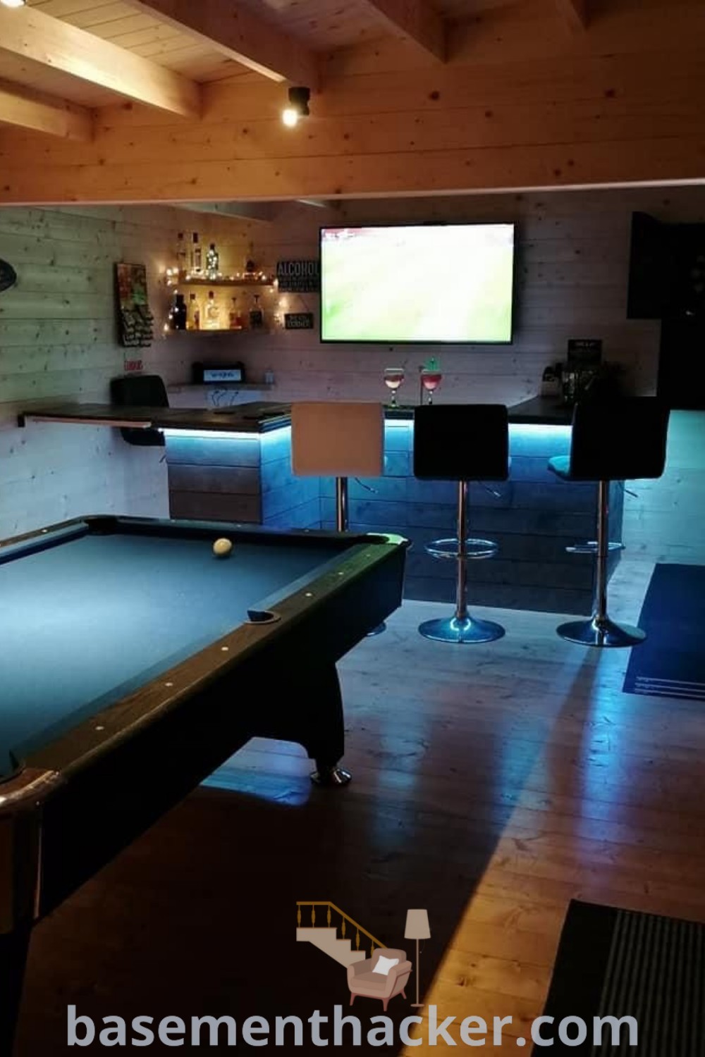 A stylish man cave with a pool table at the center, surrounded by stools and featuring a flat-screen TV on the wall. This pool room encapsulates innovative ideas for a stylish bonus space, making it perfect for entertaining. For more man cave inspirations, visit basementhacker.com.