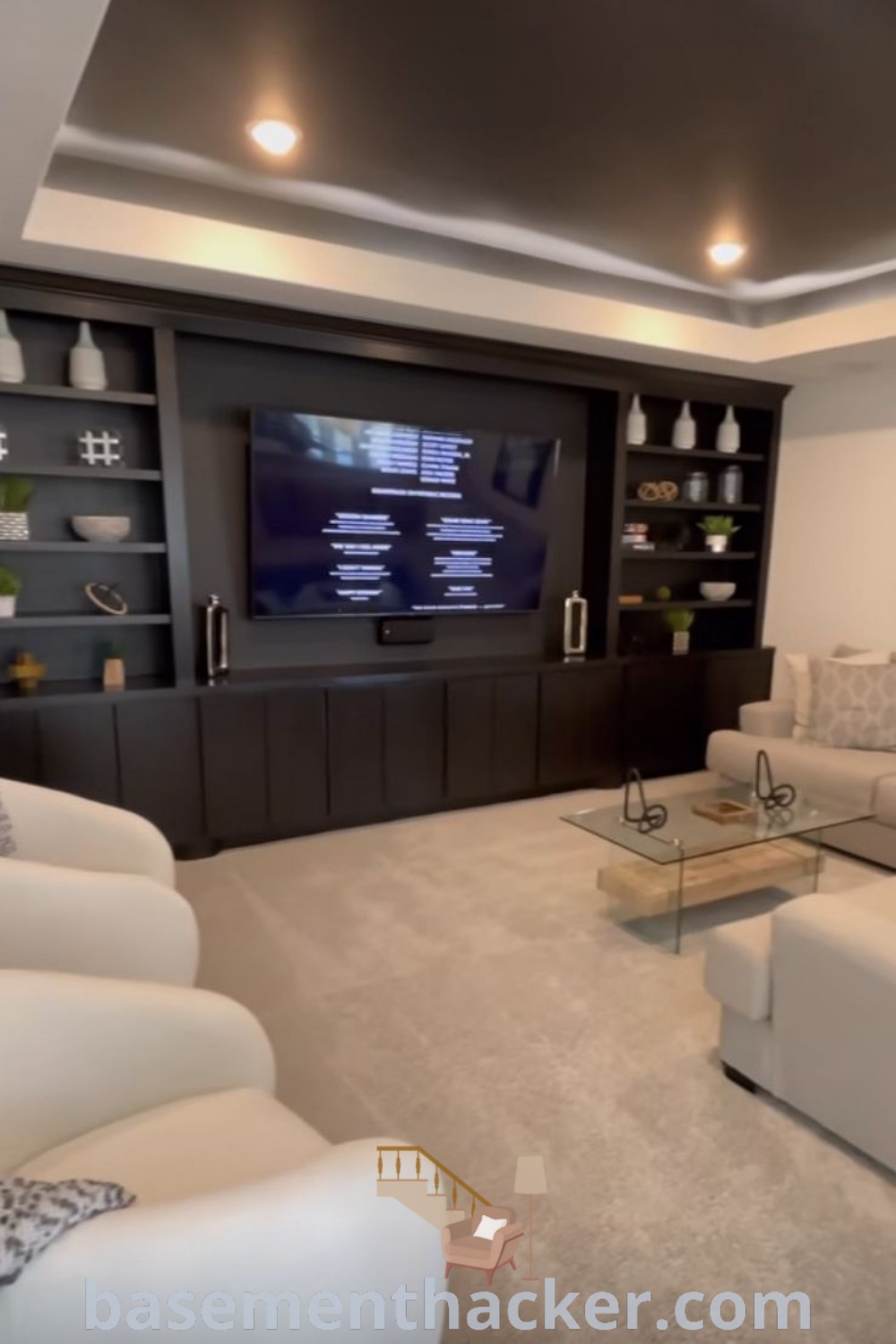 A cozy living room in a basement with furniture and an 85 inch TV mounted on the wall, showcasing stylish basement entertainment room ideas from basementhacker.com. Features floating shelves, dark grey decor, and a layout ideal for a media playroom or moody movie room experience. Perfect for family enjoyment and entertainment space transformation.