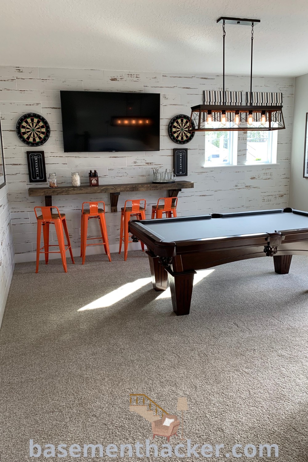 A modern pool table sits in a stylish living room adjacent to orange chairs and a flat-screen TV, showcasing innovative ideas for a stylish and functional home design on basementhacker.com. This dream basement serves as a perfect hangout room and incorporates elements from the best basement remodeling ideas for your home.