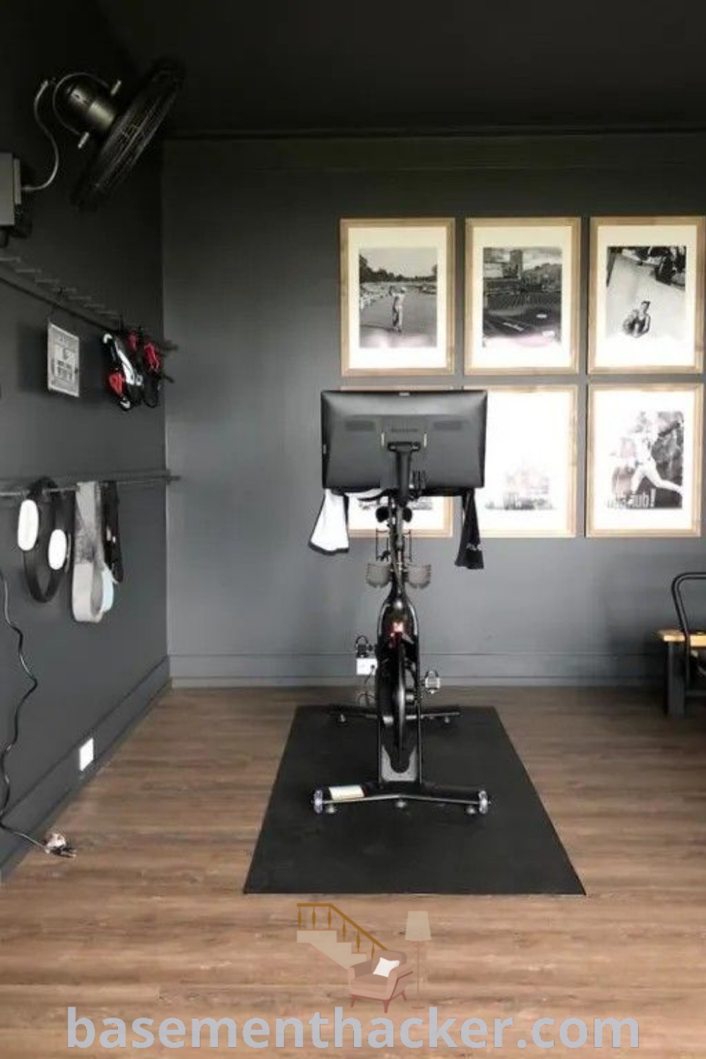 A stylish black home gym featuring a bike in the center with inspirational pictures on the walls, embodying black workout room ideas and dark workout room aesthetics. Perfect for innovative ideas for designing your home gym, this setup inspires with its sleek and functional design. Discover more home decor ideas at basementhacker.com.