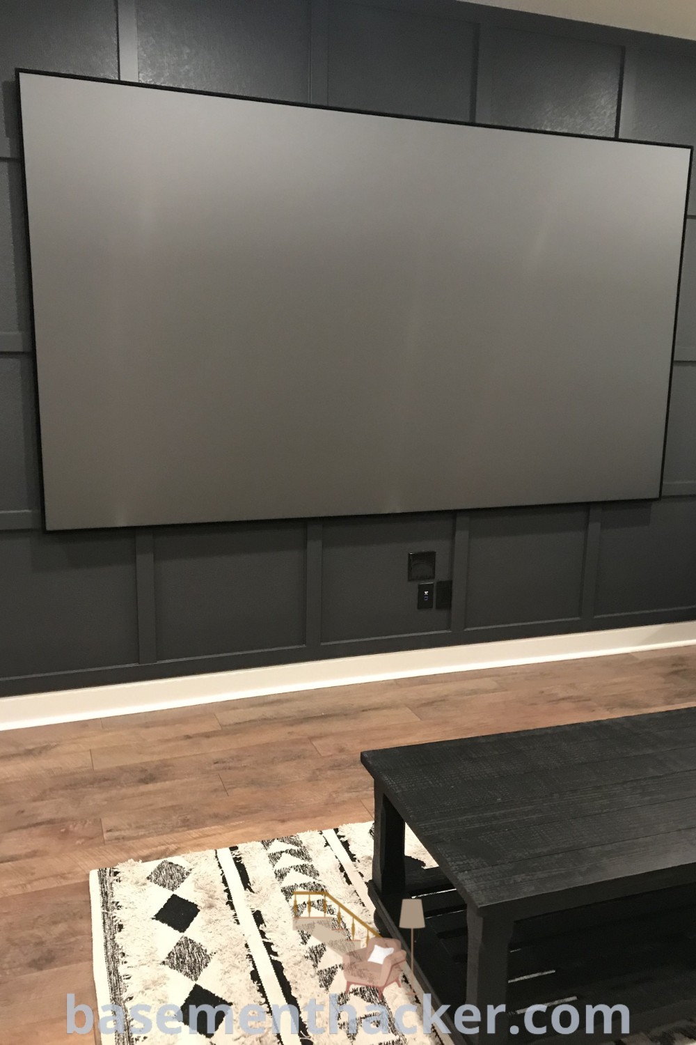 A flat screen TV mounted on the side wall of a living room, complemented by a coffee table, showcases stylish basement movie room ideas. This innovative concept includes elements like black movie room ideas and board and batten design, ideal for a cozy and organized space for ultimate entertaining at basementhacker.com.