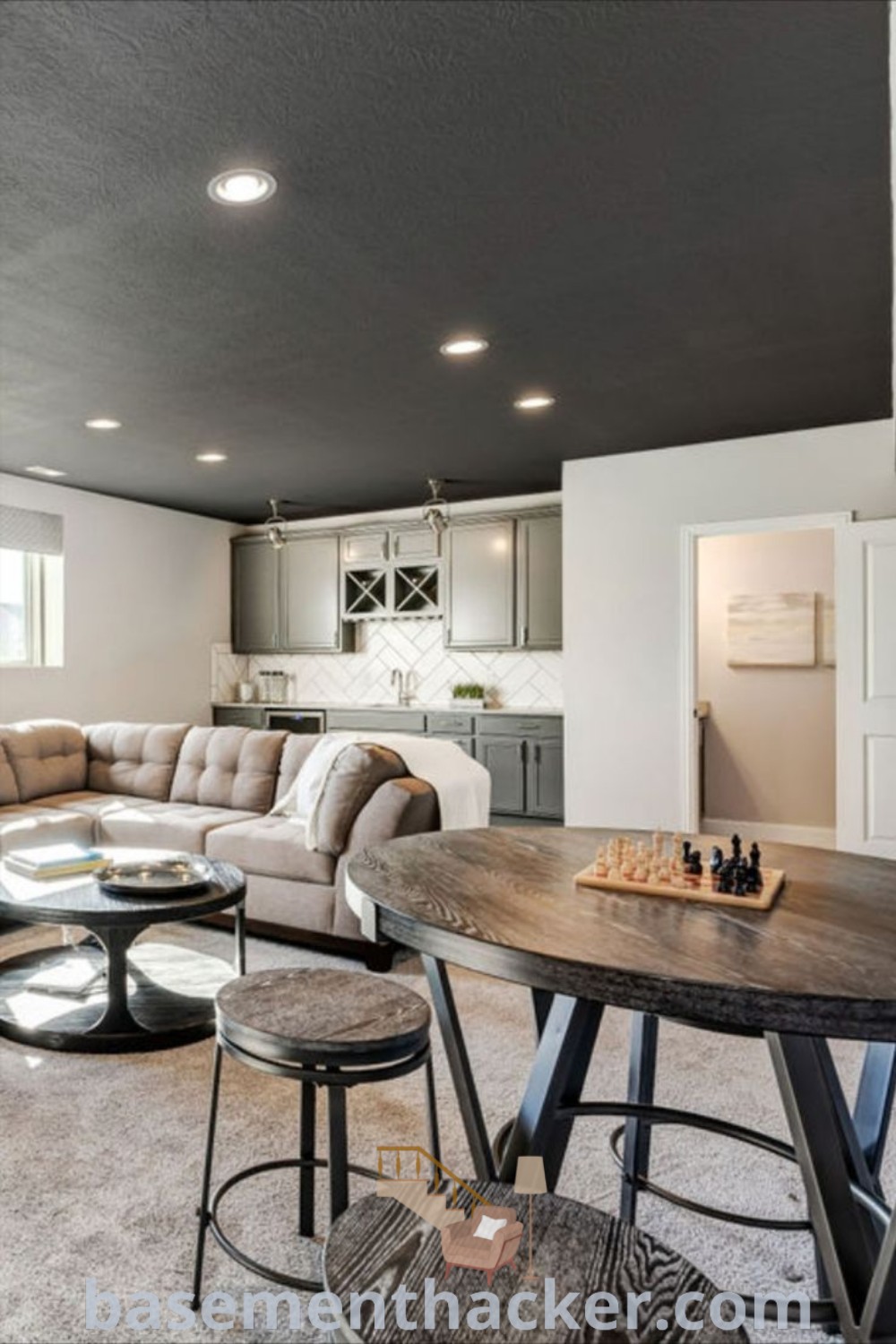 A cozy adult game room with furniture adjacent to a kitchen and dining table, designed for innovative ideas for an organized home. This stylish bonus space is perfect for family fun and entertaining, showcasing the best basement remodeling ideas. For more inspirations, visit basementhacker.com.